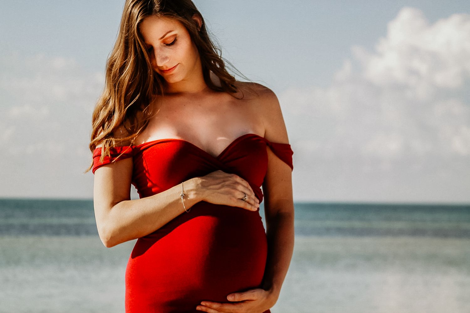 Expectant mother in red off-shoulder dress poses on beach with ocean view.