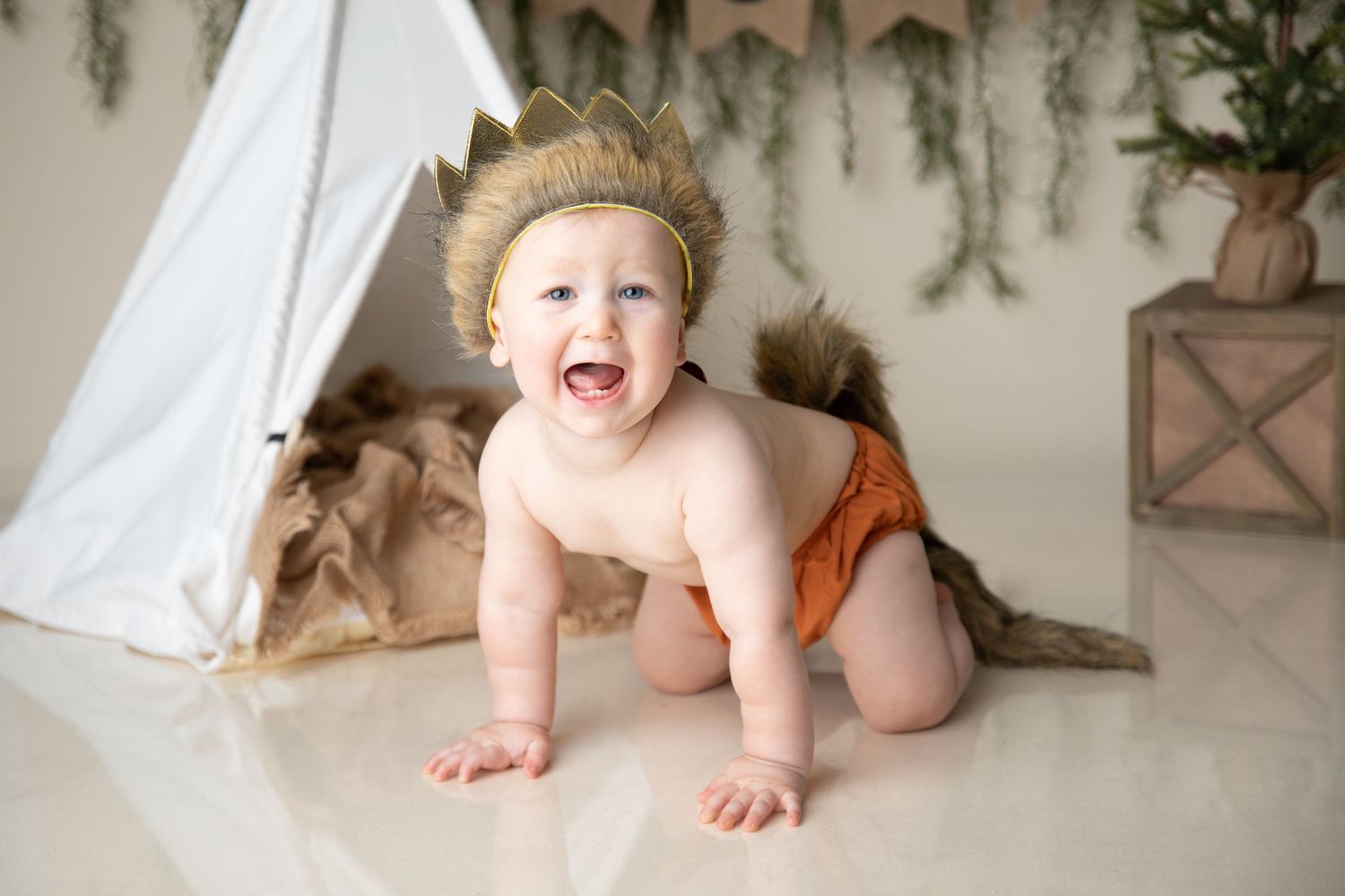 Baby wearing crown and brown diaper crawling near teepee tent setup.