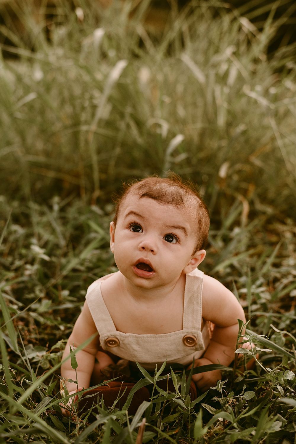 Baby in beige overalls crawling through lush green grass during a summer photoshoot.