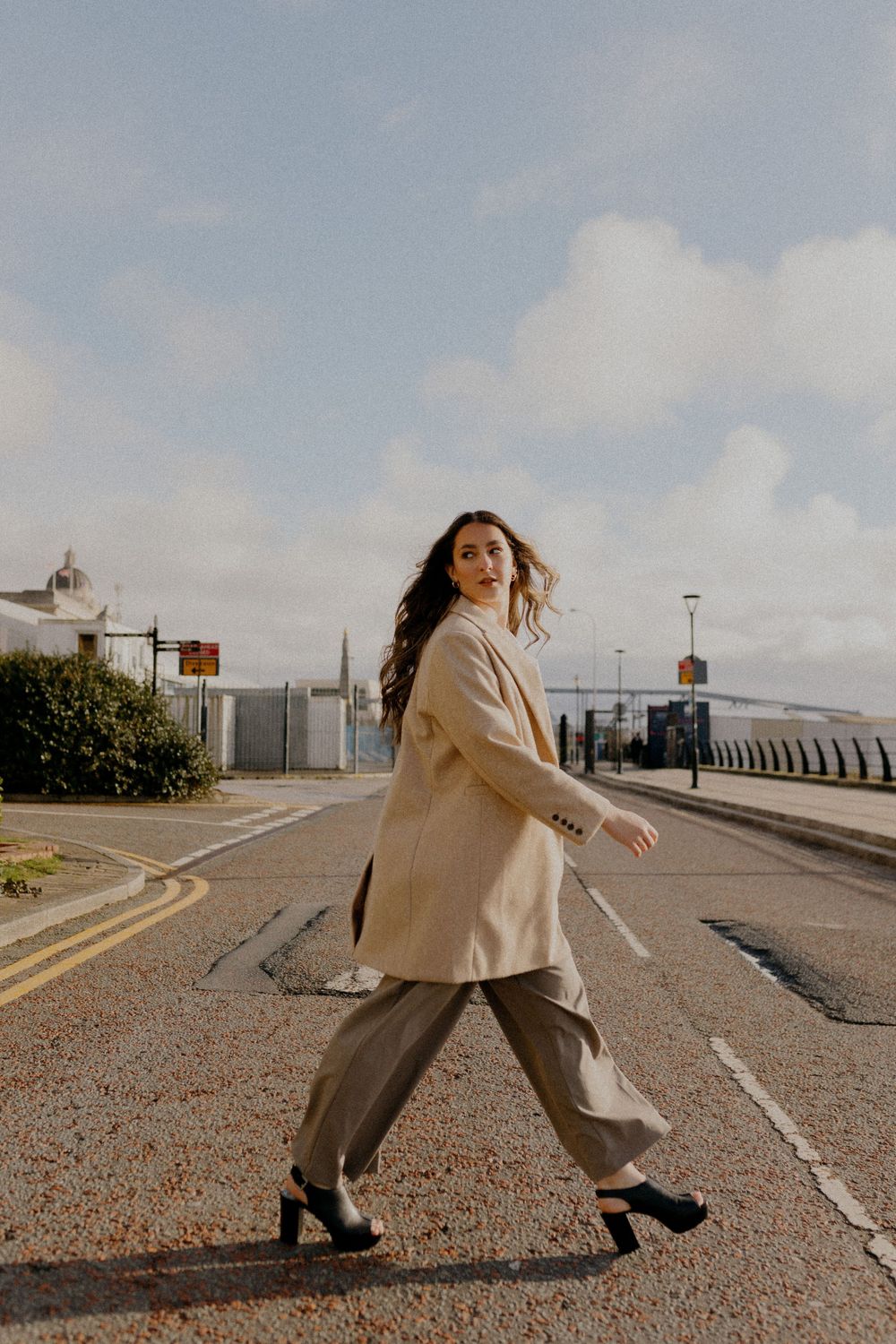 A person in a beige coat and black pants poses dramatically on an empty road.