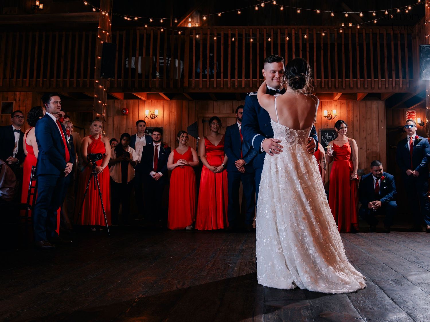 First dance moment in a rustic barn venue with red-dressed attendants watching.