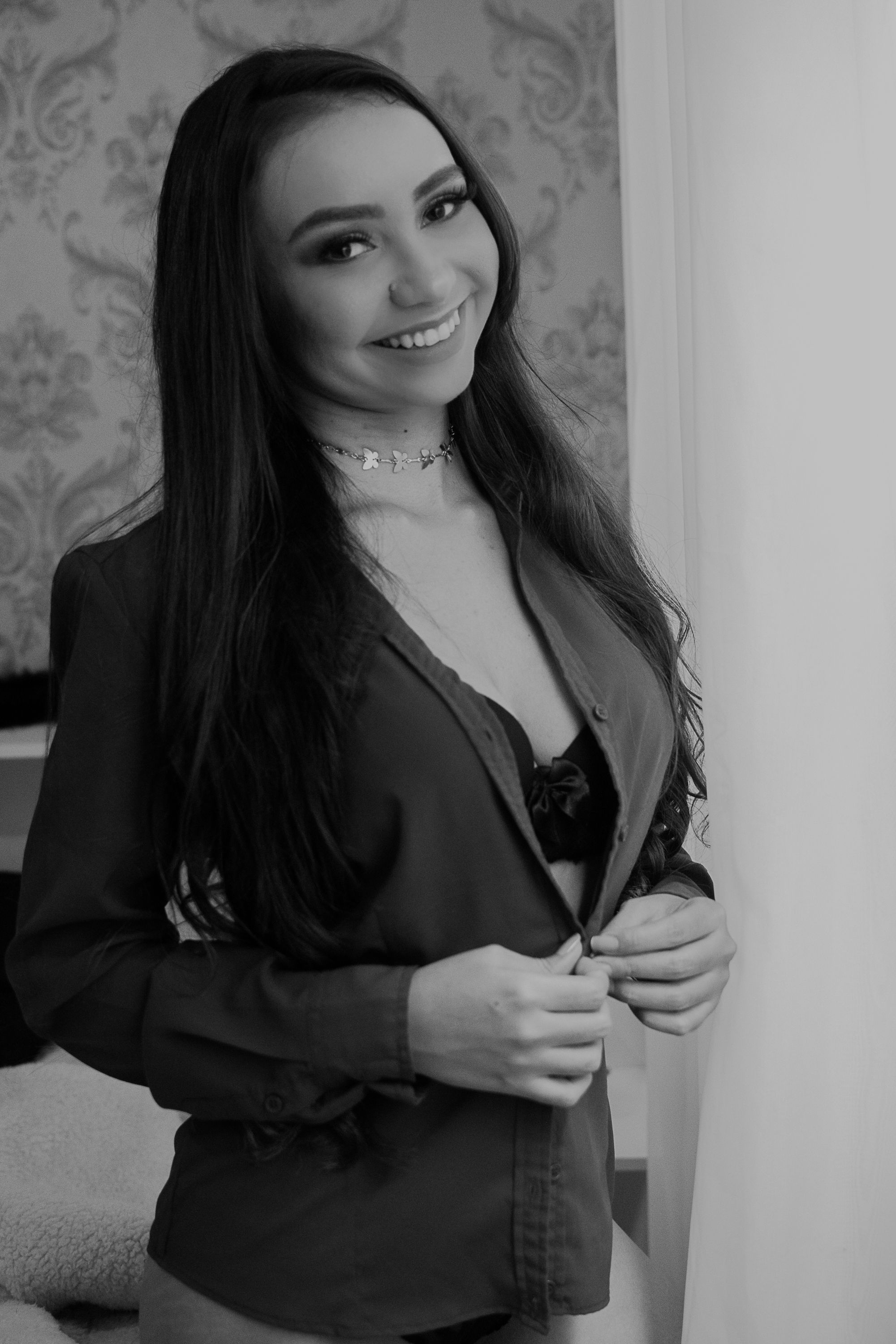 Woman in black blazer poses flirtatiously against wallpapered background.