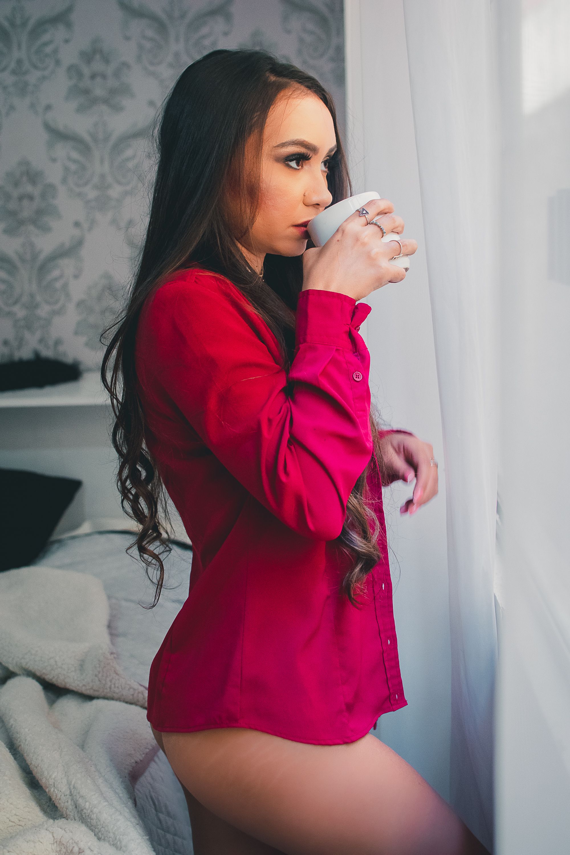 Model in vibrant red sweater poses seductively near window with damask wallpaper.