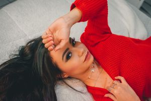 Model in vivid red knit sweater poses on white bedding with natural lighting.