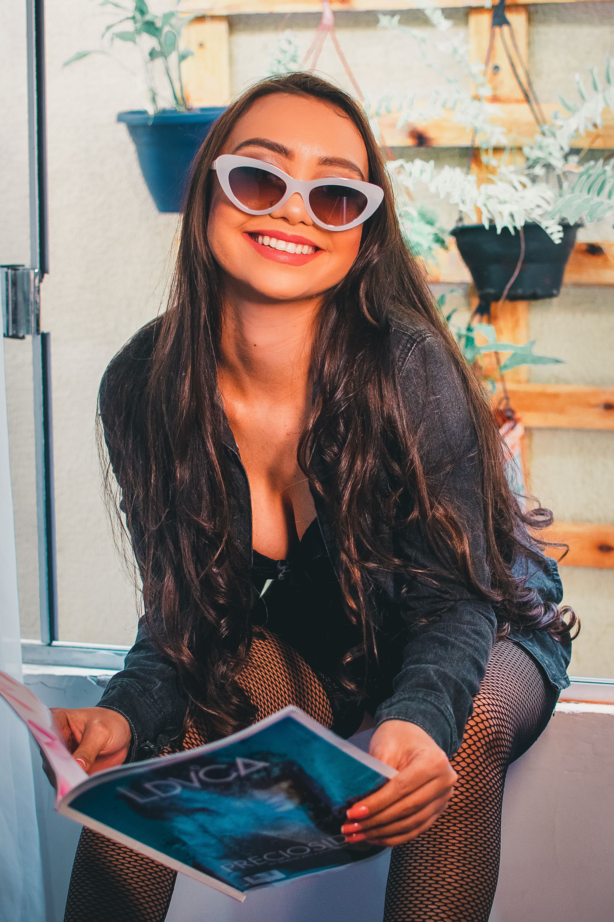 Stylish outfit with white cat-eye sunglasses and book against plant wall backdrop.