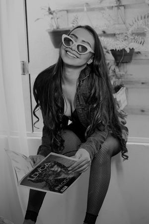 Casual reading pose in black outfit and white sunglasses against rustic wall decor.