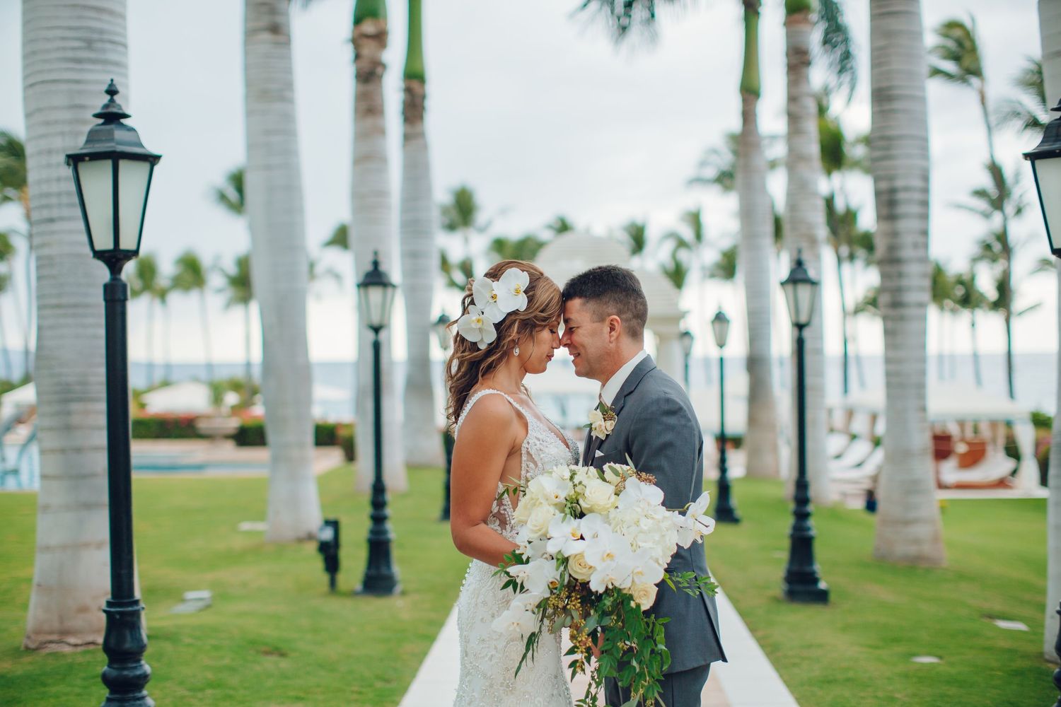 Bride and groom embrace among tropical palm trees with ocean view at sunset.