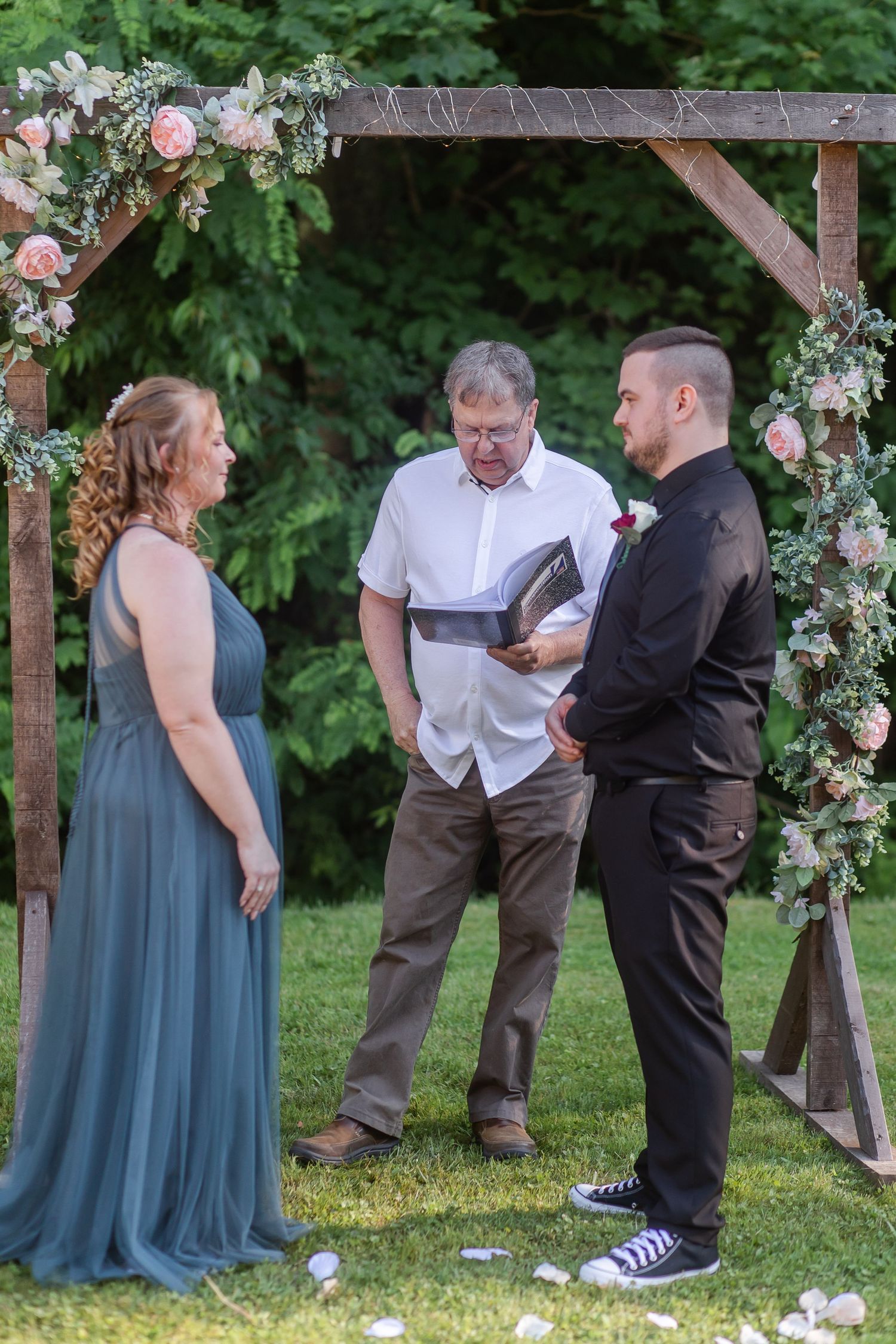 Wedding party gathered under a wooden arch decorated with pink and white flowers in a garden setting.