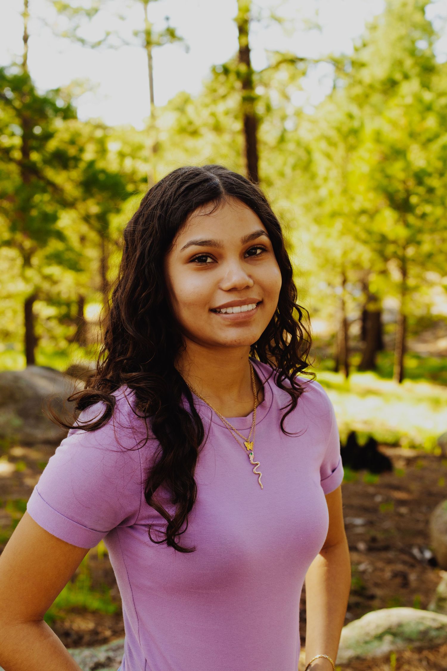 Person in pink t-shirt with long dark hair poses outdoors in natural setting.