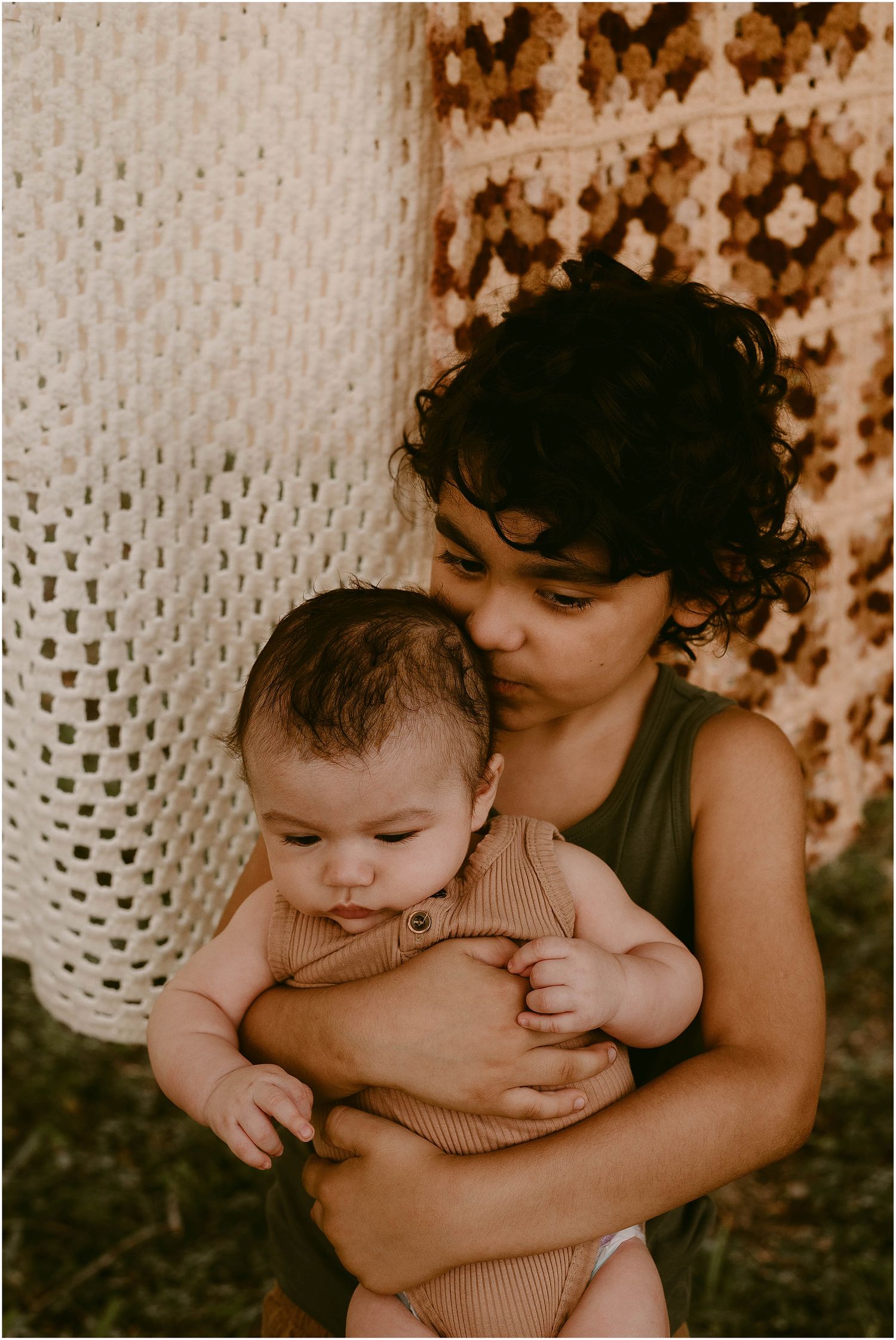 A tender moment captured in sepia tones showing an embrace against a backdrop of white crochet and brown floral patterns.