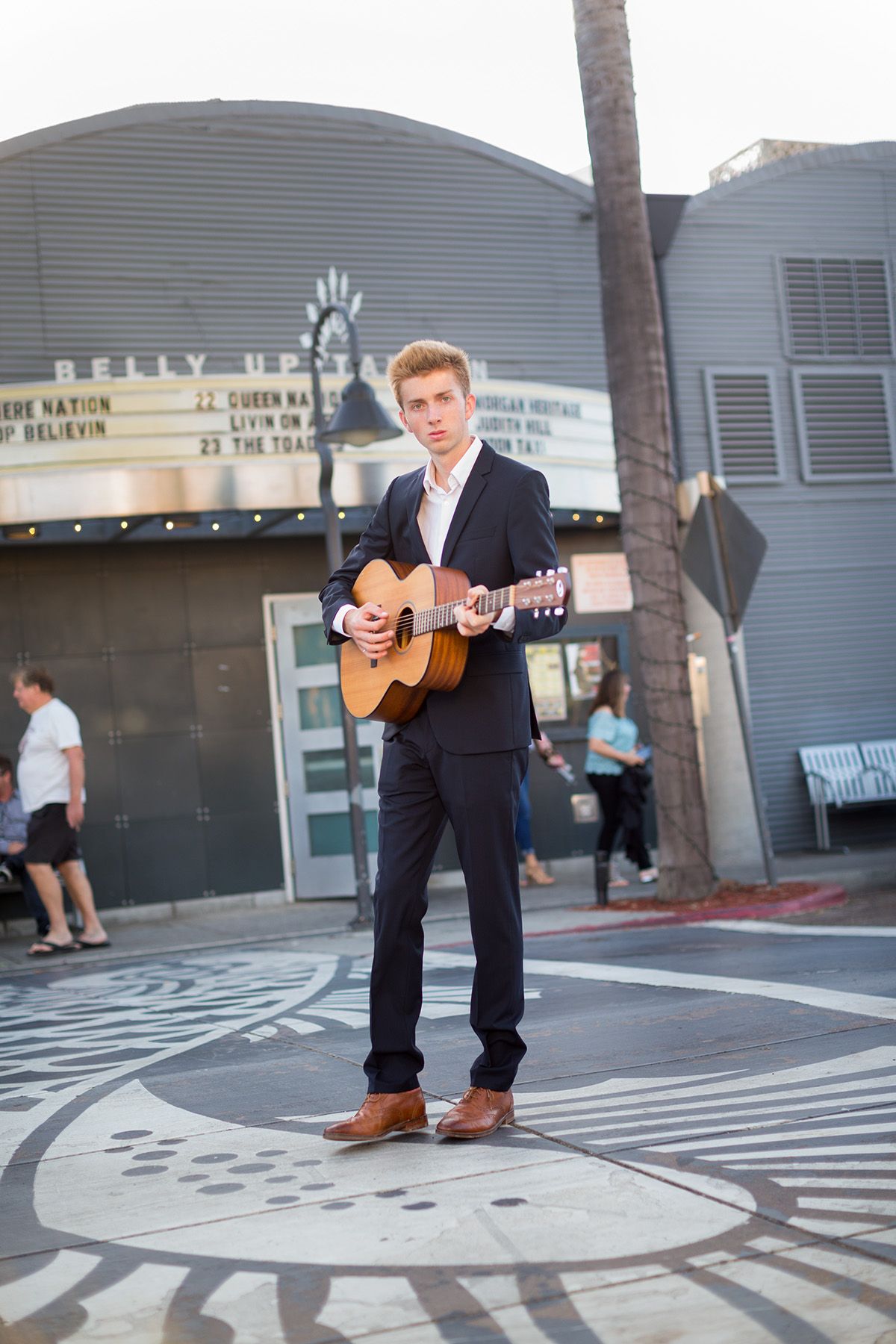 Musician playing acoustic guitar outside a theater wearing a formal black suit.