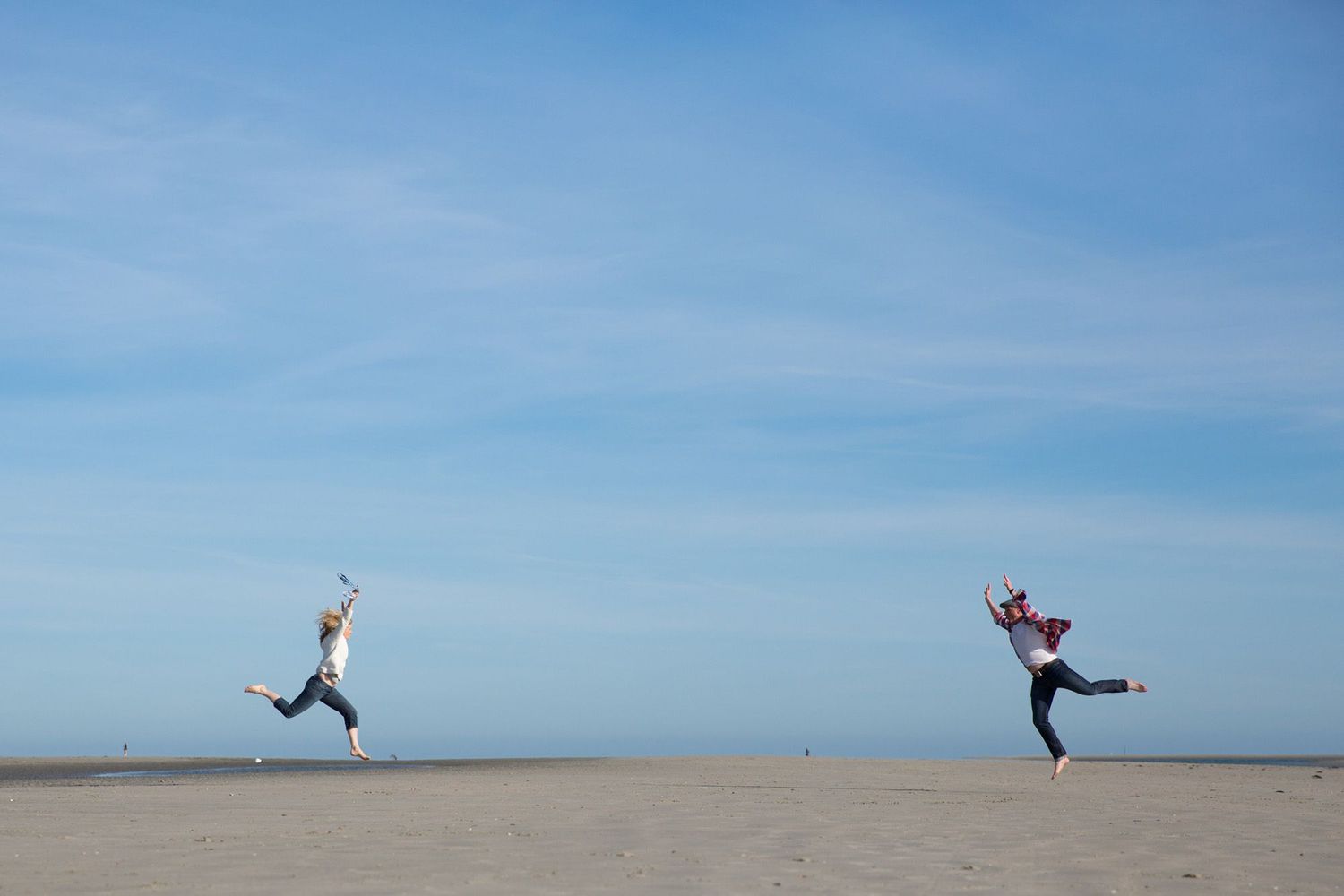 Two people joyfully jumping in the air on an empty beach with clear blue skies.
