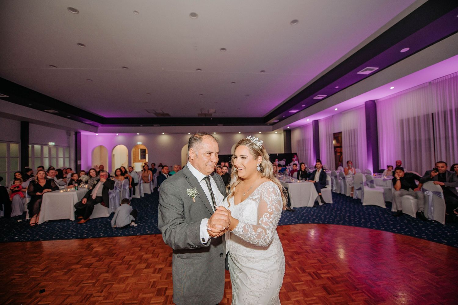 Wedding couple dances in decorated reception hall with purple lighting and white table settings.
