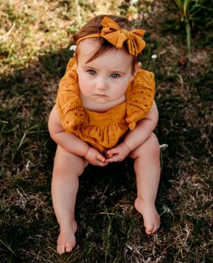 A baby wearing a mustard yellow dress and matching bow sits on grass outdoors.