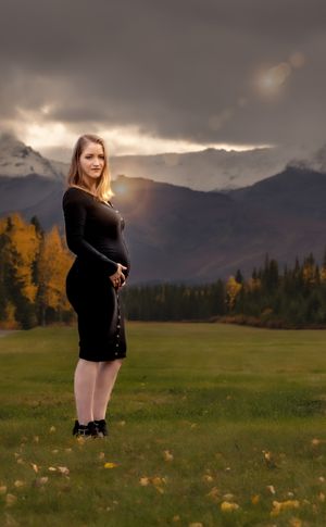 A person poses in a fitted black dress against a backdrop of mountains and dramatic sunset clouds.