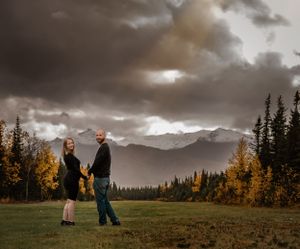 A couple holds hands while standing in a meadow with dramatic mountain views and autumn colors.