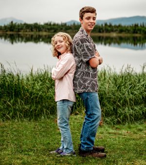 Two siblings pose back-to-back by a tranquil lake with mountains visible in the distance.