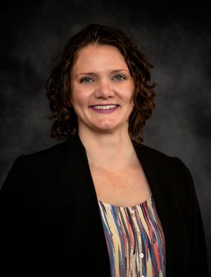 Professional headshot of a person in black blazer and colorful striped top against dark background.
