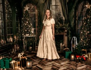 Woman in vintage cream dress poses amid Christmas decor with wrapped presents and lanterns on herringbone floor.