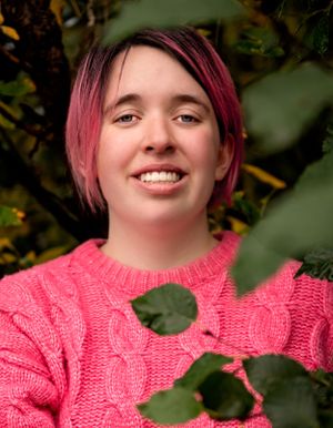 Person with pink hair wearing coral sweater smiles warmly against green foliage.