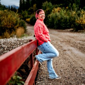 Person in pink top leans against red railing on dirt path with autumn foliage in background.