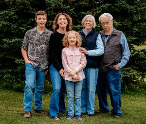 Family members stand together on grass with evergreen trees forming a scenic backdrop.