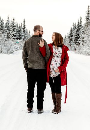 Couple in winter attire share an intimate moment on a snowy path surrounded by evergreen trees.