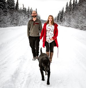 Couple walking their black dog on a snowy winter trail through evergreen forest.