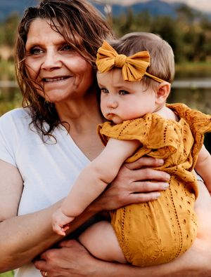 A tender outdoor portrait of an adult holding a baby in a mustard yellow dress with matching bow.