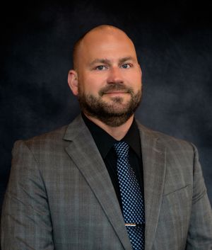 Professional headshot of a person in a gray suit and blue tie against dark background.