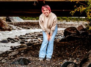 Person in beige sweater stands among rocks by flowing stream.