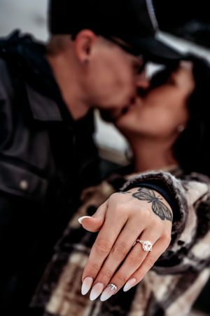 Close-up of an engagement ring on a manicured hand with white nails and tattoo details.