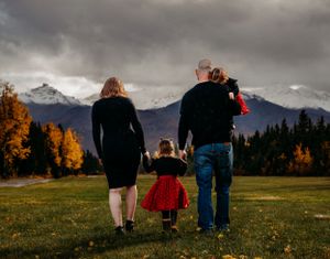 A family of three walks together towards mountain views with golden fall foliage surrounding them.
