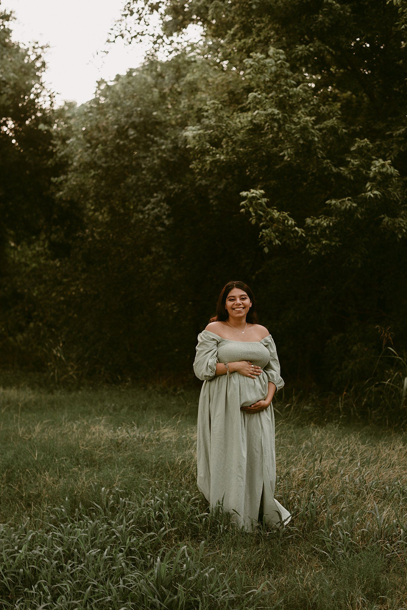 Maternity photo of woman in flowing grey dress standing in meadow at sunset.