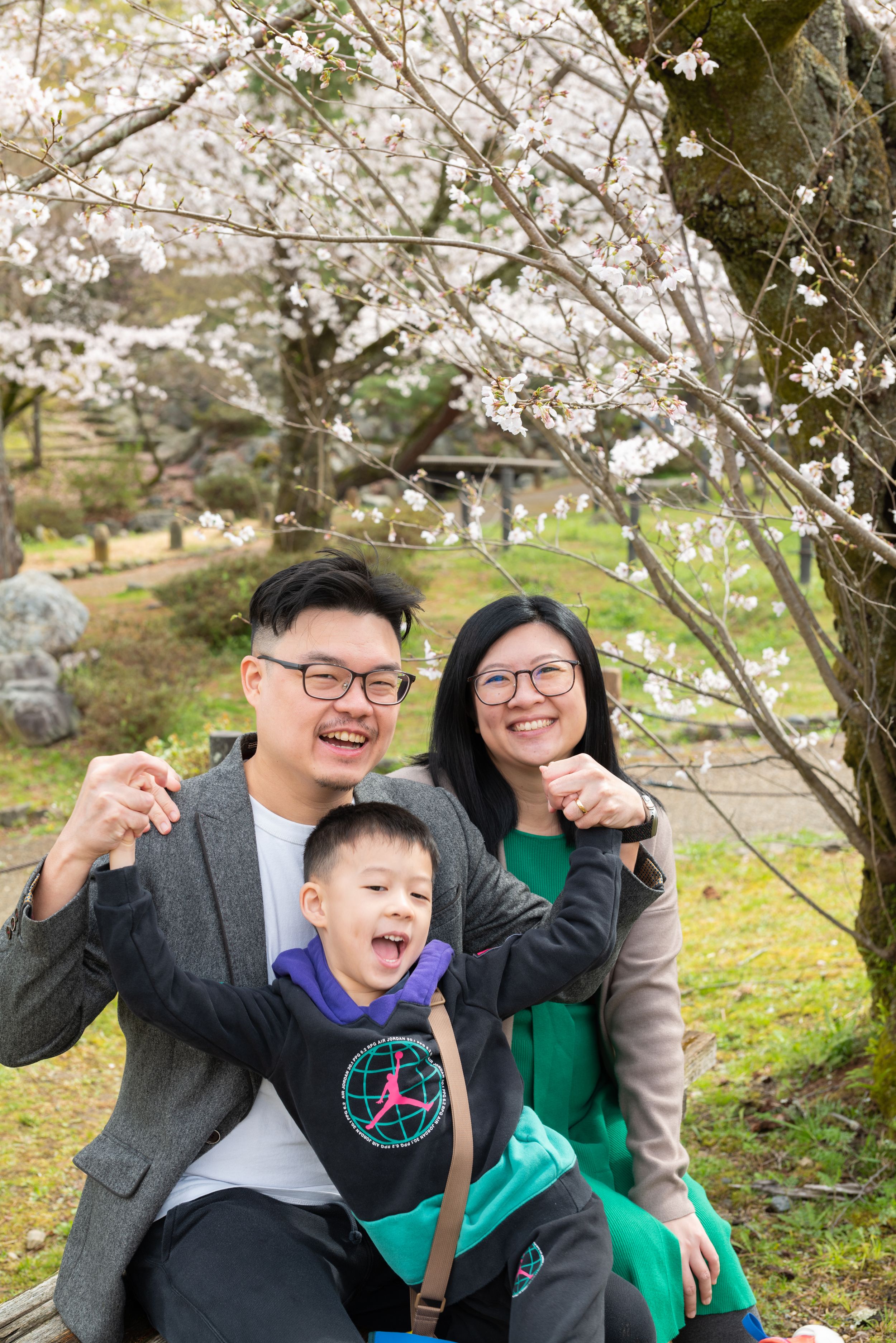 Joyful Asian family making fun poses under white cherry blossom trees in garden.
