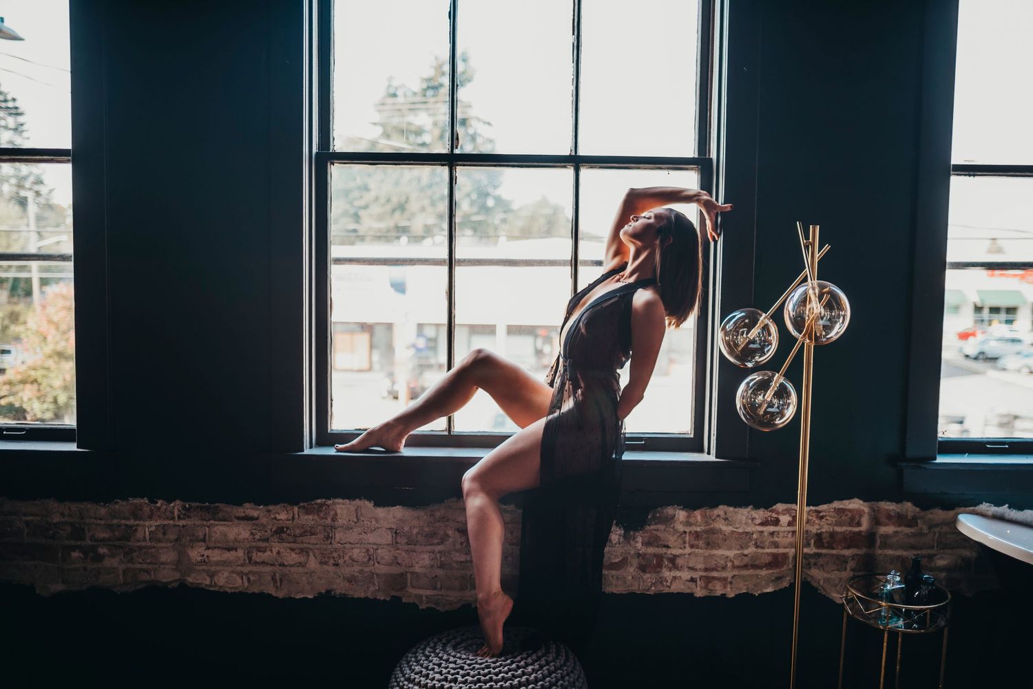 Woman in black lingerie posing gracefully by a large window, showcasing her confidence and sensuality in soft lighting.