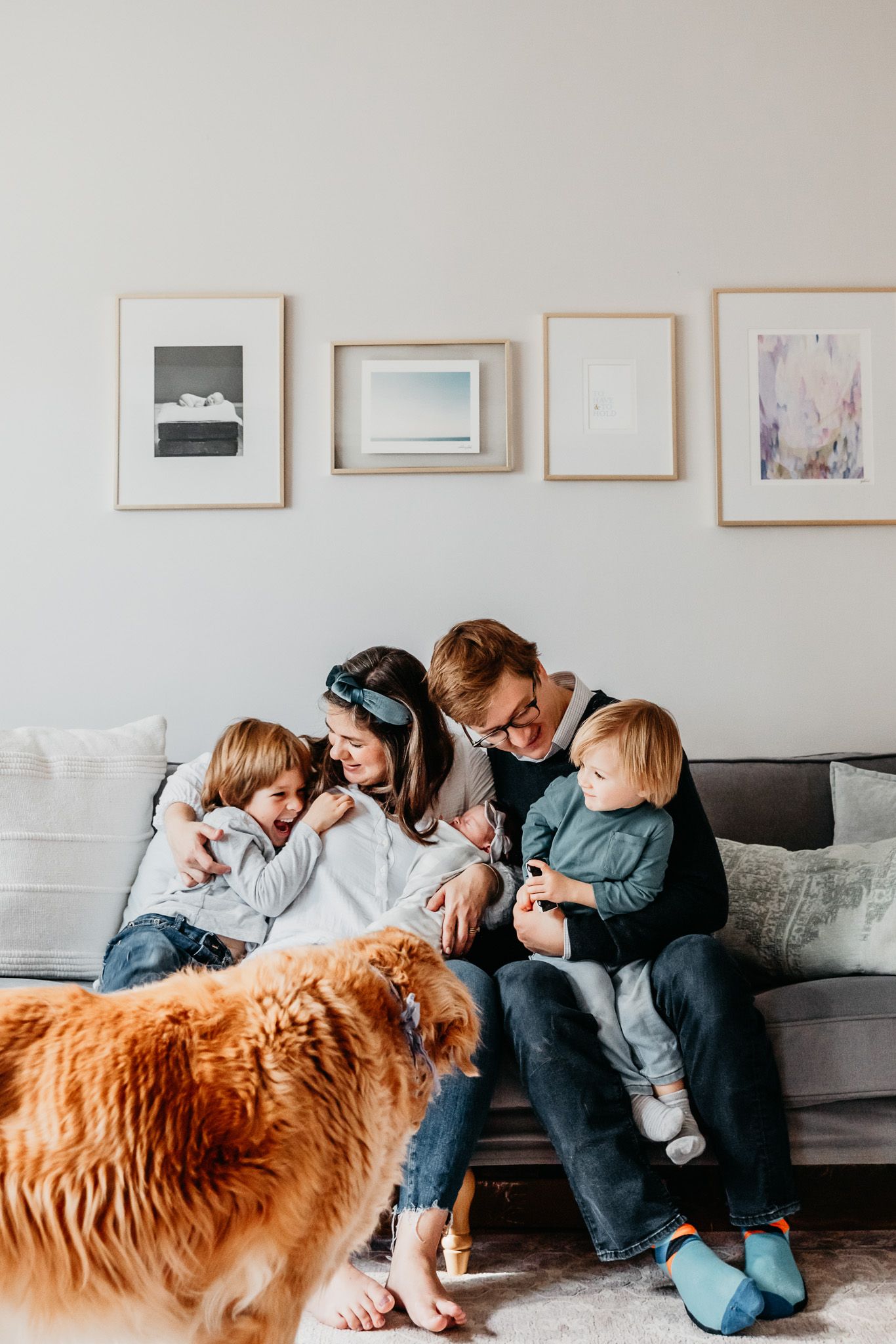 A family laughs together in their living room. Mom holds onto the newborn baby who is surrounded by her family and dog.