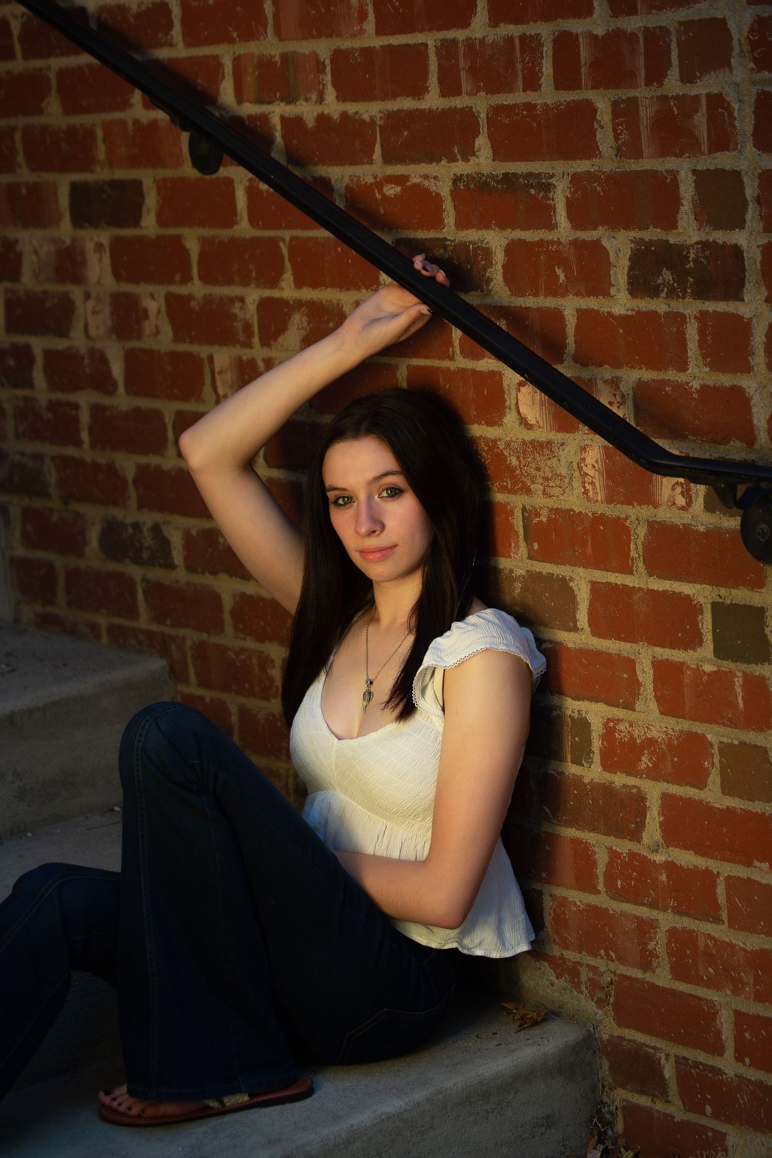 Person in white top sits on concrete steps against brick wall with black handrail.