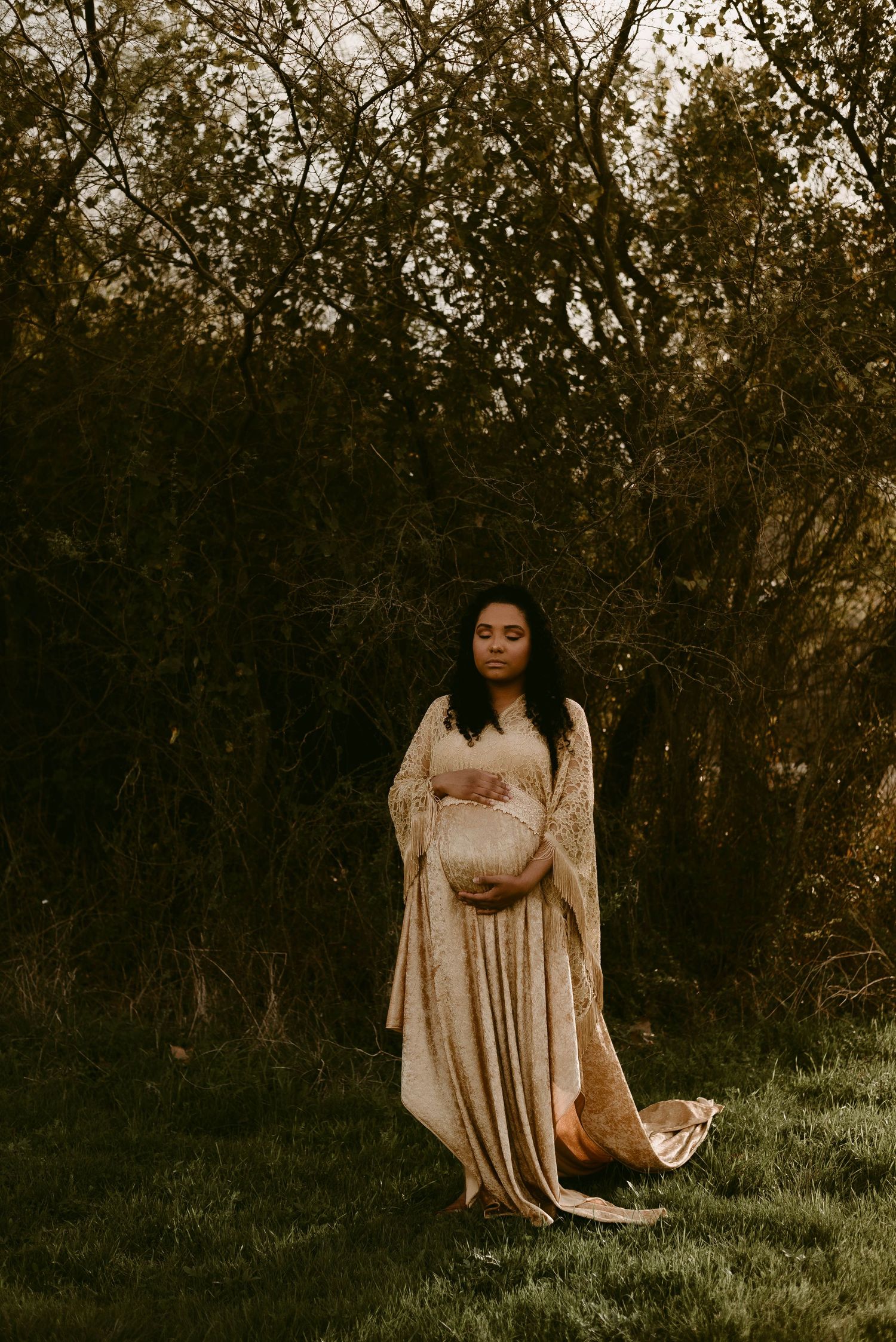 Full-length maternity portrait in cream gown against dark forest background.
