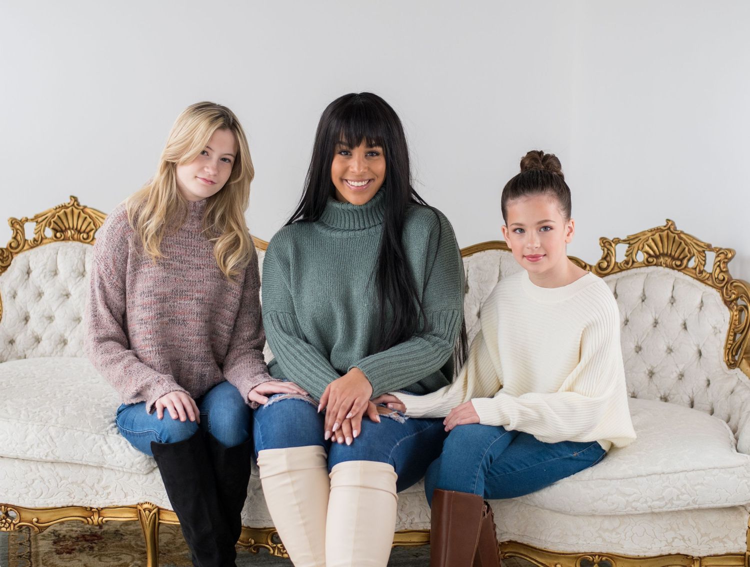 Three women in cozy sweaters sit together on an elegant vintage couch.