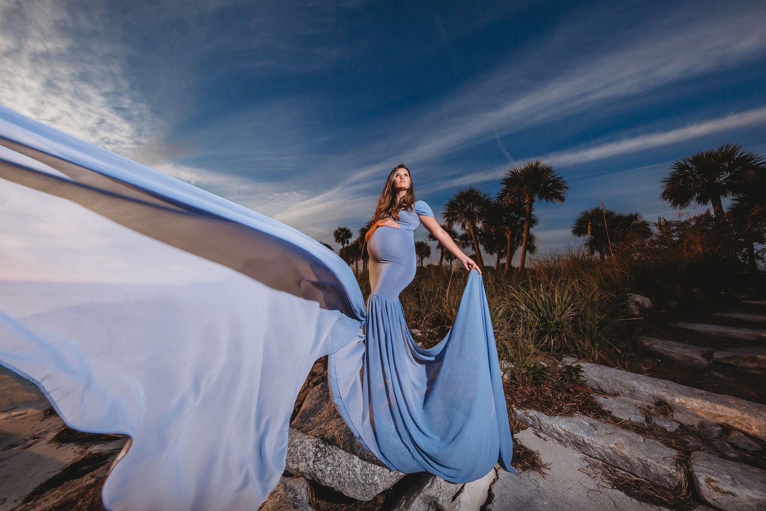 Artistic portrait with billowing light blue fabric against coastal landscape at dusk.
