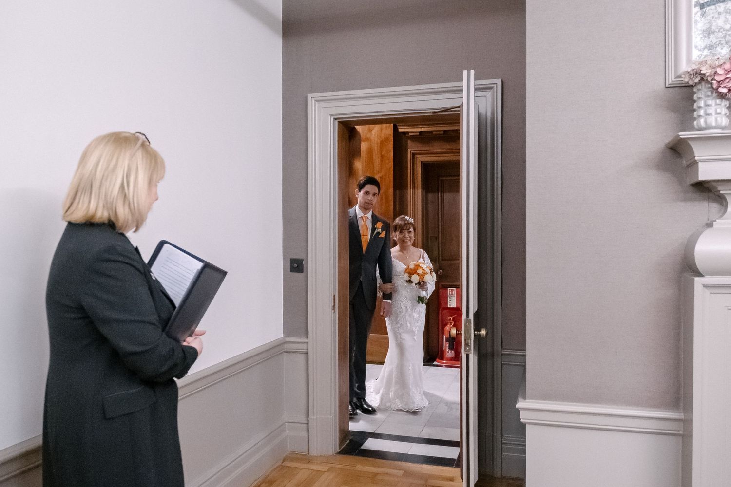 The bride enters the room with her escort as the registrar looks towards them.
