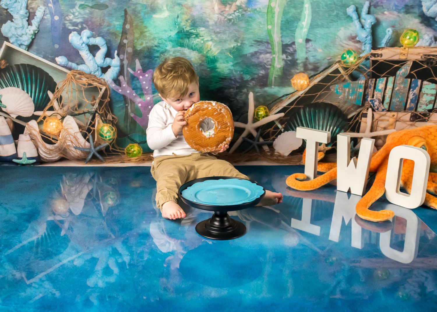 Toddler boy sitting on reflective blue floor holding pretzel at underwater themed birthday party.