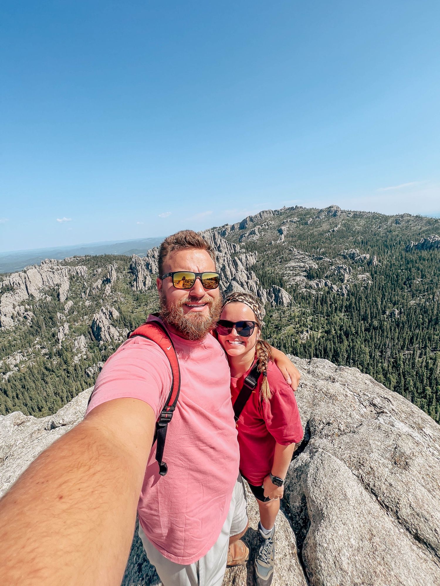 Two hikers take a selfie from a scenic mountain overlook with rocky cliffs and forested valley visible in background.