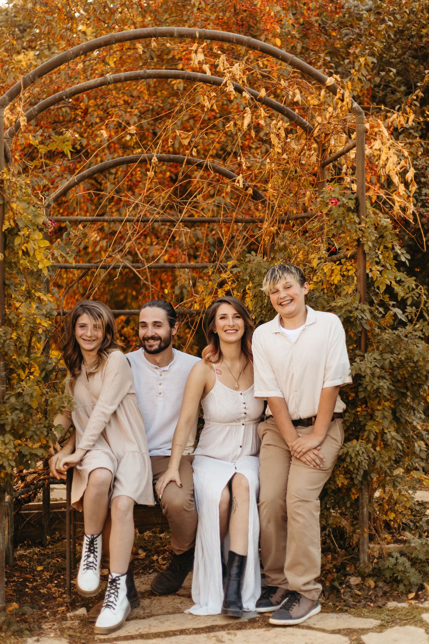 Family group in coordinated neutral outfits sitting together on wooden bench against fall foliage.
