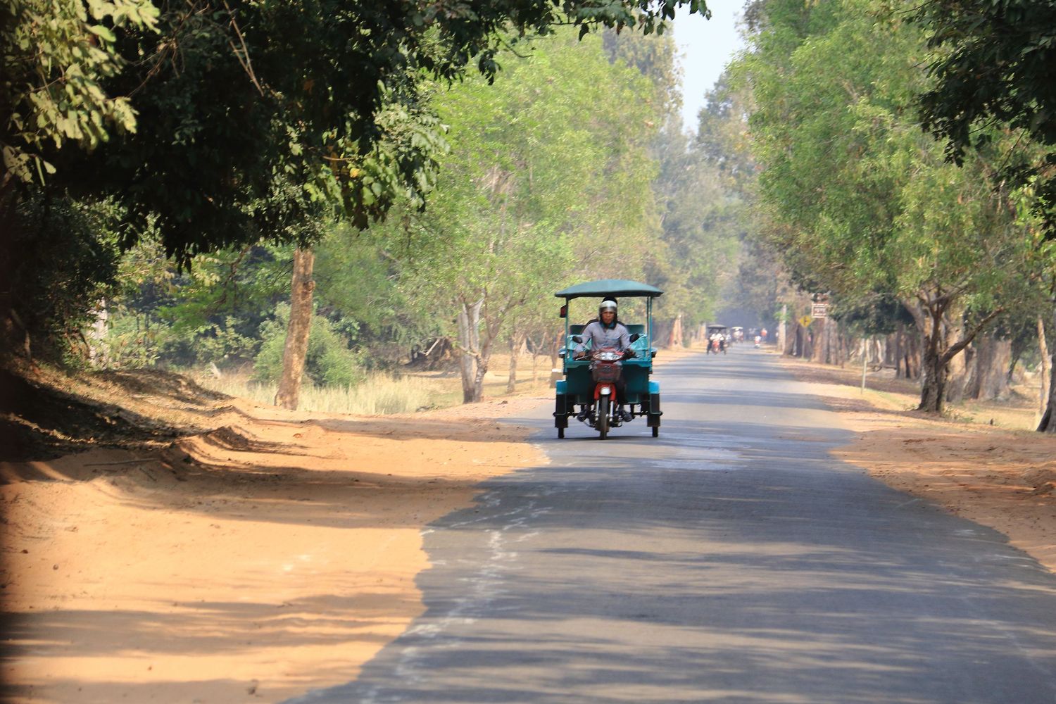 Tuk tuk driving down a tree-lined rural road in Cambodia.
