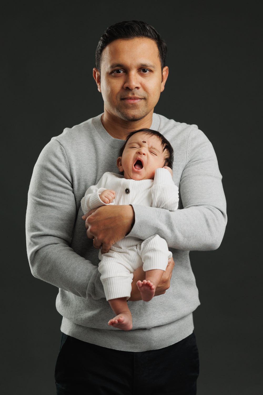 A parent in a grey sweater holds a yawning newborn baby wearing white against a dark background.