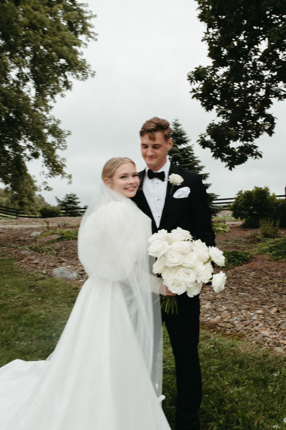 A wedding couple poses outdoors under trees in formal attire with white flowers.