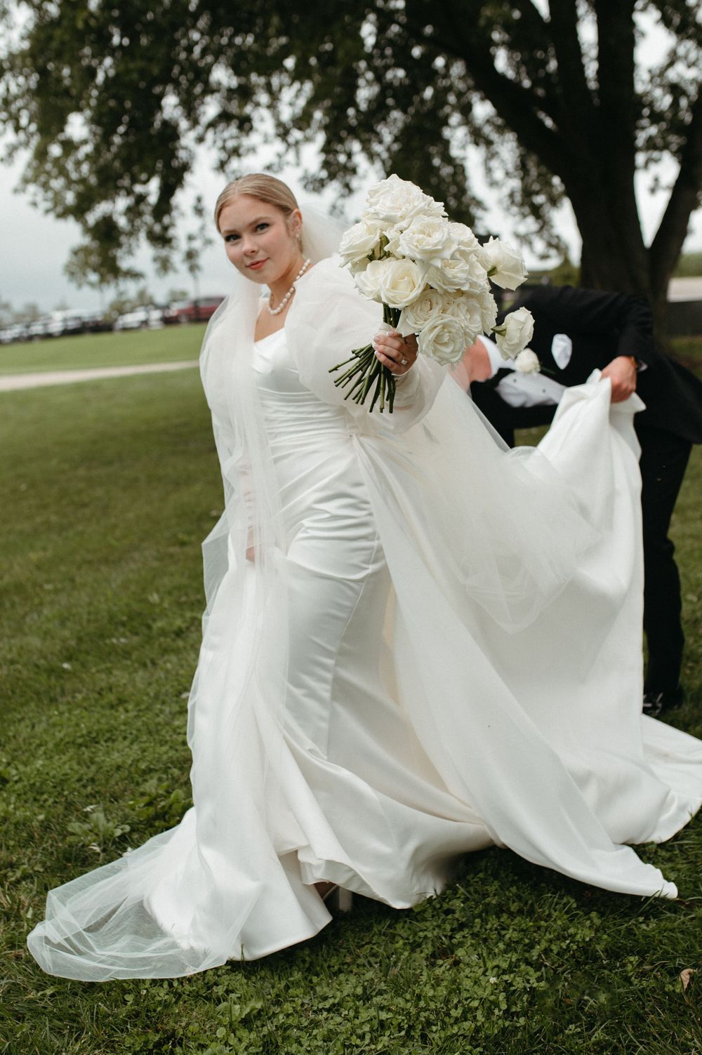 A bride in an elegant white wedding gown holding a bouquet of white roses outdoors.
