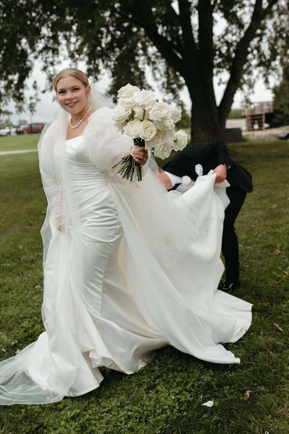 A bride in a fitted white gown with flowing train poses against a backdrop of trees.
